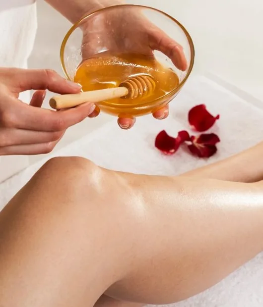 Waxing scene: hand holding a glass bowl of warm amber wax and a wooden spatula over a leg, rose petals on a towel