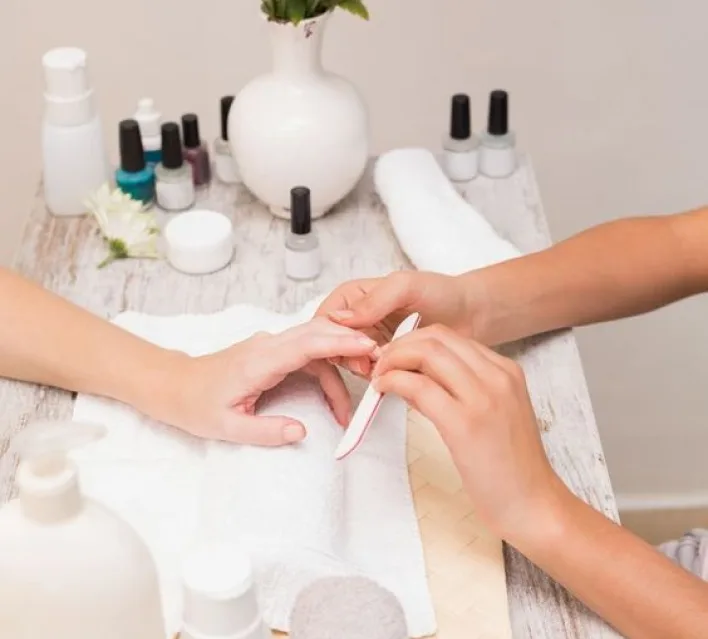 Nail technician filing and shaping a client's nails at a flower-topped table with polish bottles and a white vase in the salon