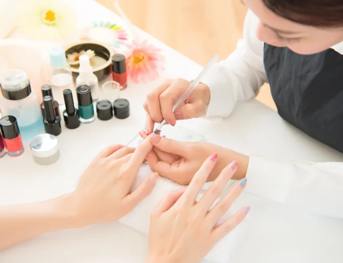 Manicurist painting a client's nails at a table with colourful polish bottles and flowers