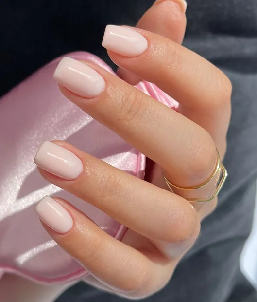 Close-up hand with milky square natural nails holding pink satin, gold ring on finger